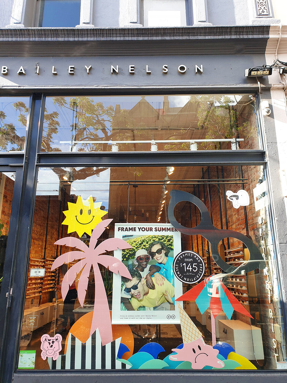 A colourful installation of cut-out shapes decorates. Bailey Nelson retail store window. The  window props include a giant pair of eyeglasses, stripy beach umbrella, sun with smiley face, black and white striped starburst shape, melted ice cream with sad 