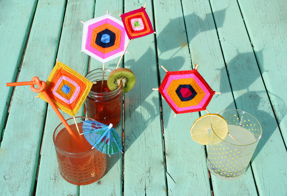 Three tropical cocktails with colourful, handmade woven yarn drink stirrers sit on a rustic green table.