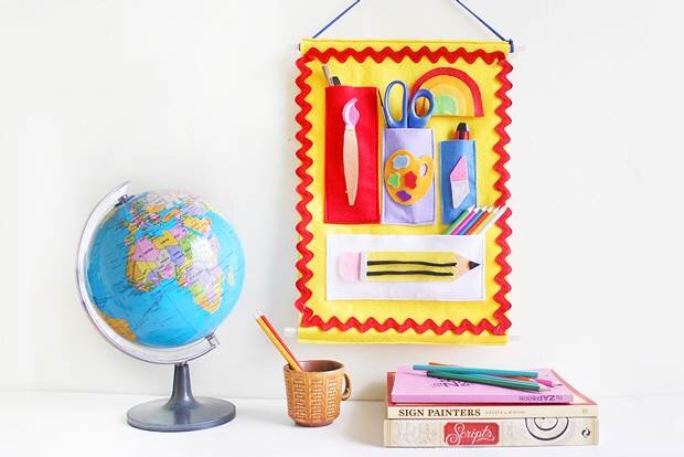 A yellow felt organiser with red tripping and coloured pockets hangs on a white wall with books, a mug and a globe